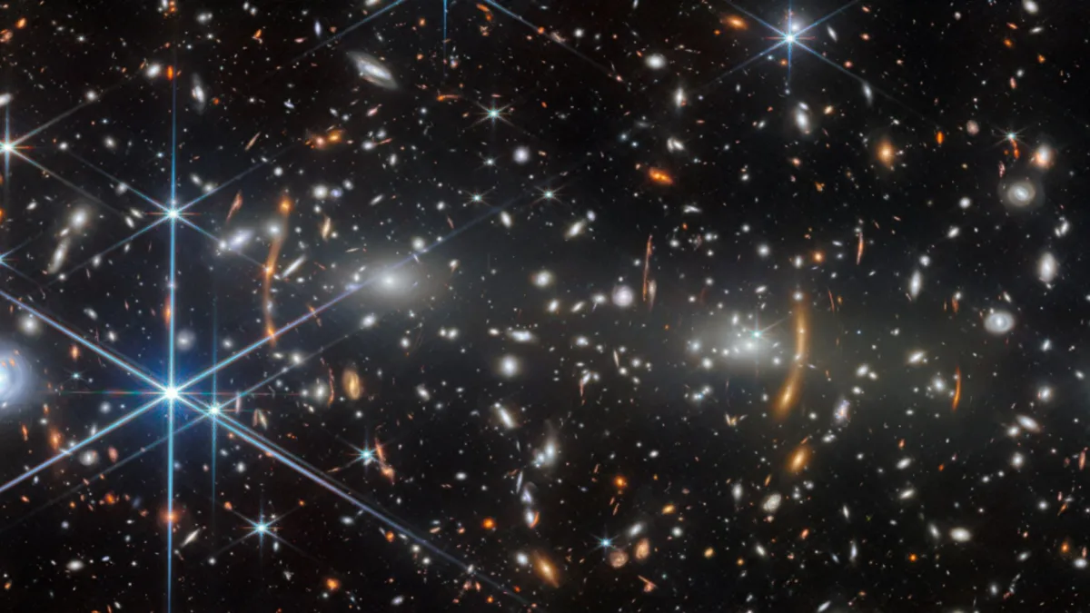 Image 24: A black space background is speckled by yellow, white, blue and orange stars including blue stars with long diffraction spikes.