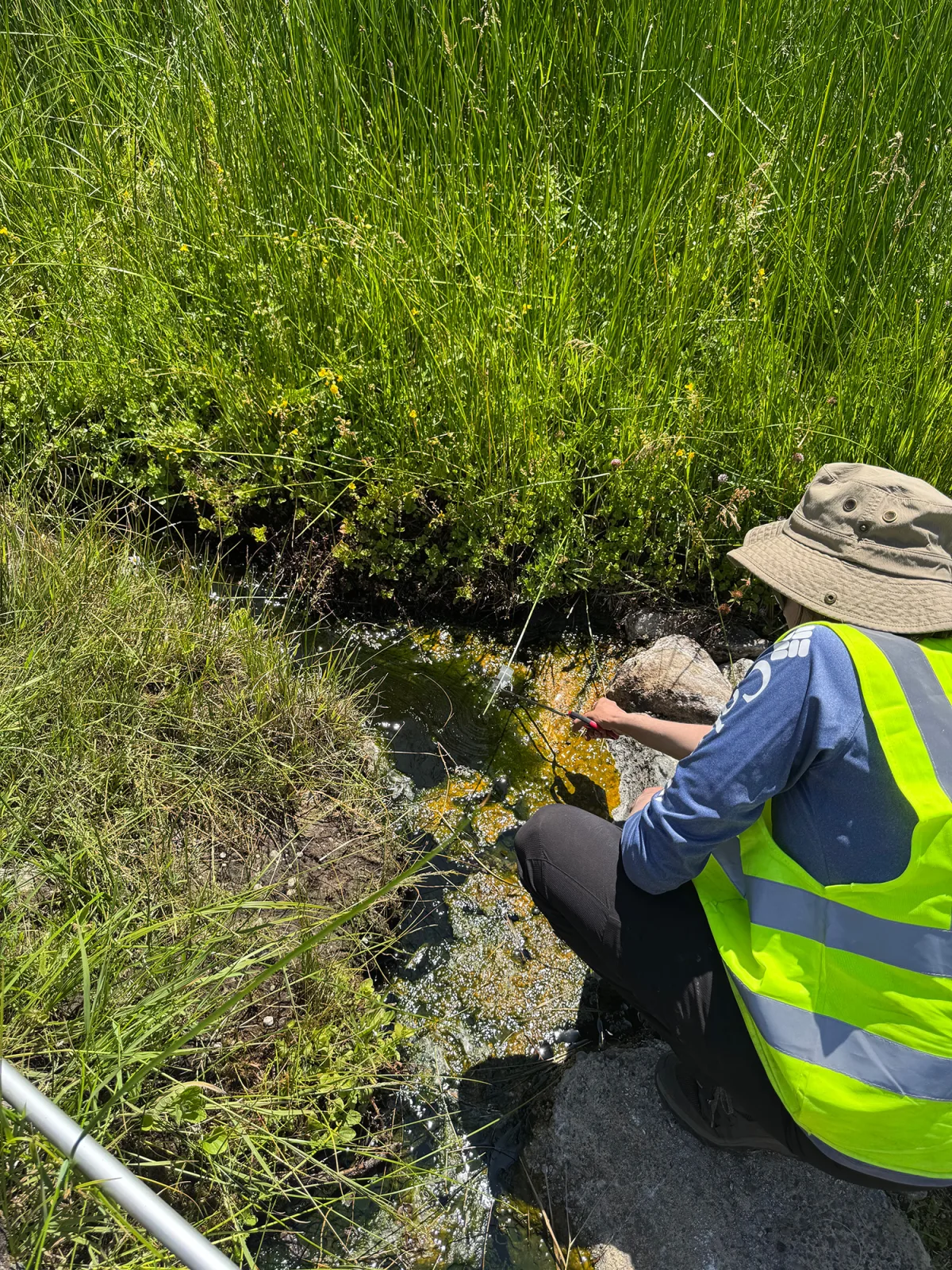 Image 41: A person wearing a yellow reflective vest and tan bucket hat leans over a small creek.