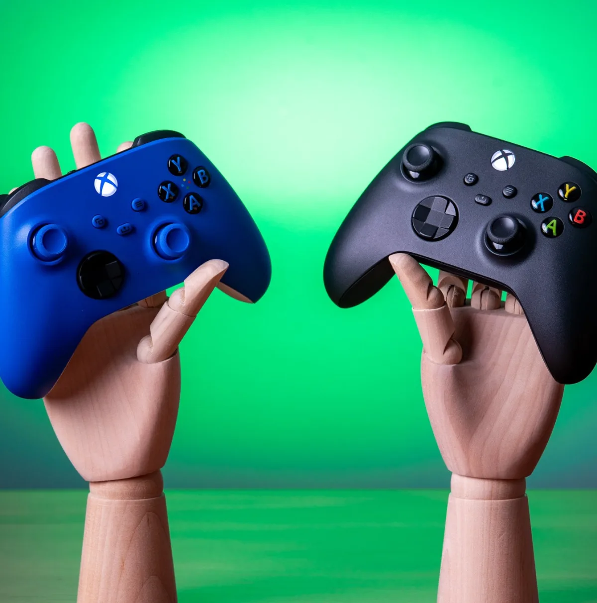 Image 7: Two Xbox controllers, one blue and one black, being held by wooden mannequin hands.